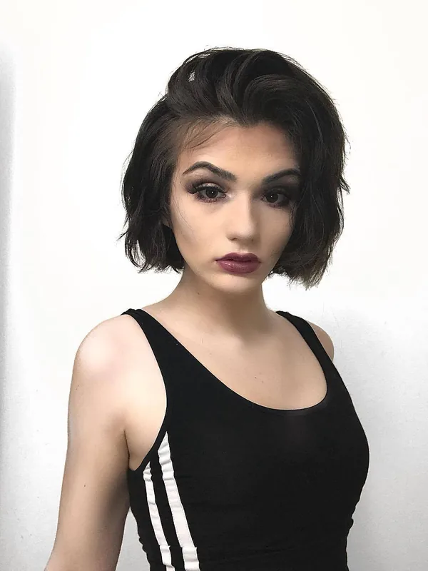 Portrait en plan rapproché d'une personne aux cheveux courts noirs, maquillée, portant un haut noir à bandes blanches, devant un fond blanc.