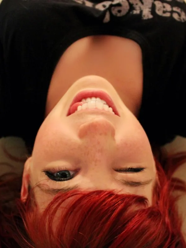 Portrait en plongée d'une personne aux cheveux rouges, souriante, allongée sur un sol carrelé, portant un t-shirt noir.