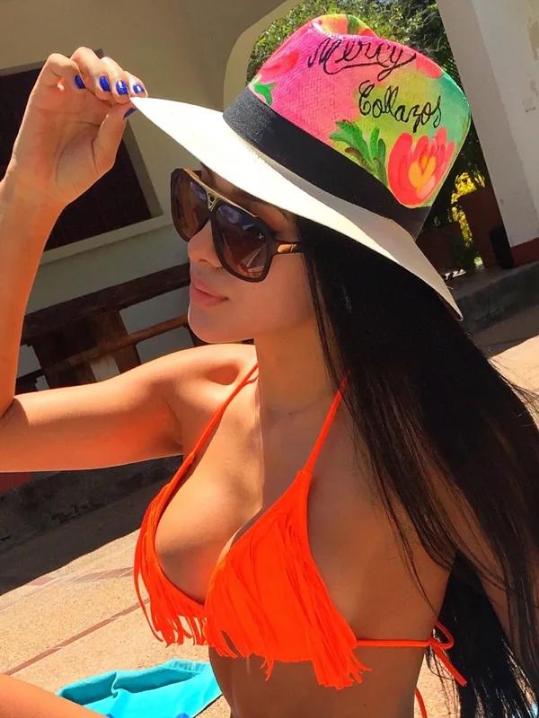 Personne en maillot orange à franges, chapeau coloré, lunettes de soleil, main levée, ensoleillé