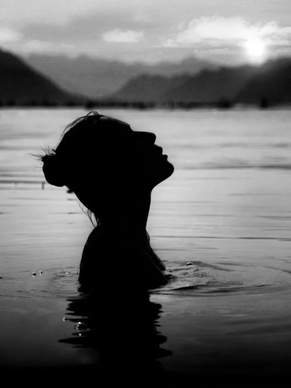 silhouette noire de personne dans l'eau, tête levée, regard vers le haut, coucher ou lever de soleil, montagnes en arrière-plan