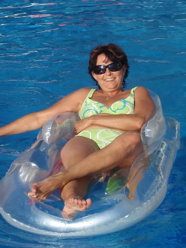Personne en maillot de bain vert et blanc sur matelas gonflable transparent dans une piscine bleue, portant lunettes de soleil