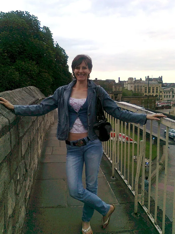 Personne souriante en veste et jean, posant sur un chemin pavé avec vue sur ville et arbres
