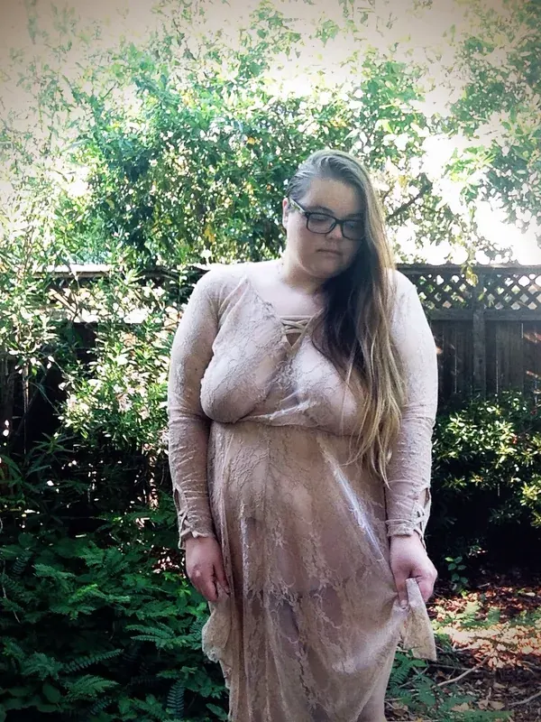 Portrait d'une personne en robe de dentelle claire, debout dans un jardin verdoyant, regardant vers le bas.