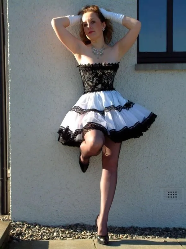 Femme en robe noire et blanche à dentelle, gants blancs, posant contre un mur extérieur en journée.
